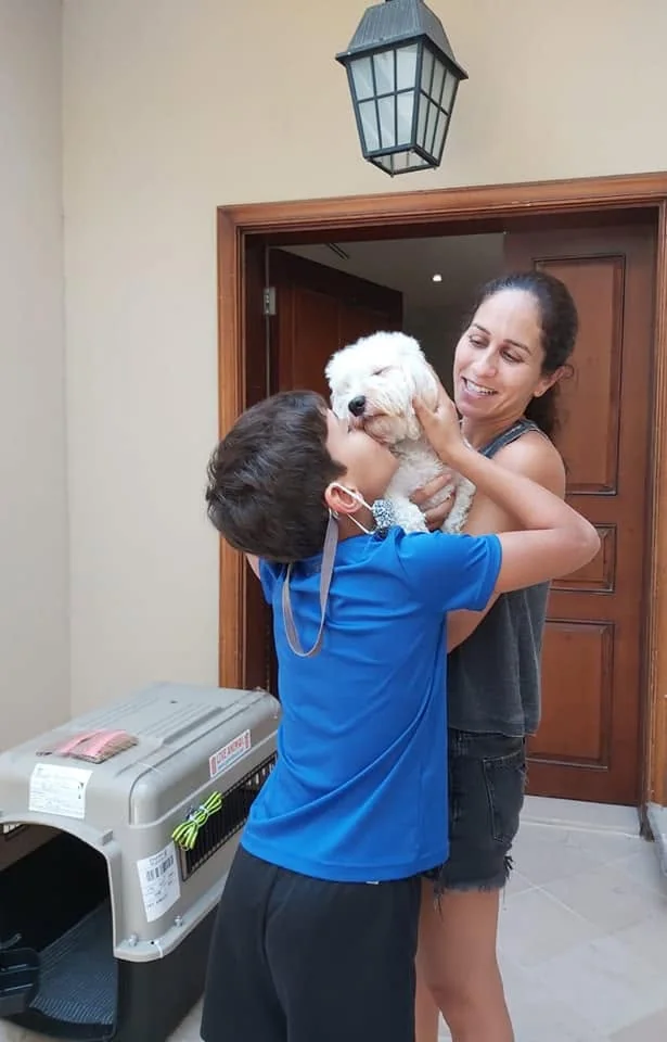 A woman and child embracing a white dog beside a pet travel crate.